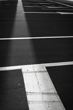 A black and white image of a parking lot with clearly defined white lines marking parking spaces. The surface appears smooth and the lines are slightly worn, suggesting regular use. There's a visible patch of light creating a contrast against the dark pavement.