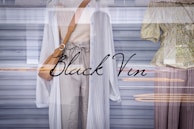 Two mannequins in a store window display are dressed in light, casual clothing. One wears a long white cardigan over a beige knitted top and light trousers, complemented by a tan shoulder bag. The other mannequin is dressed in a green floral blouse with buttons and wide brown trousers. The display reflects urban street scenery in the glass, adding layers to the image.