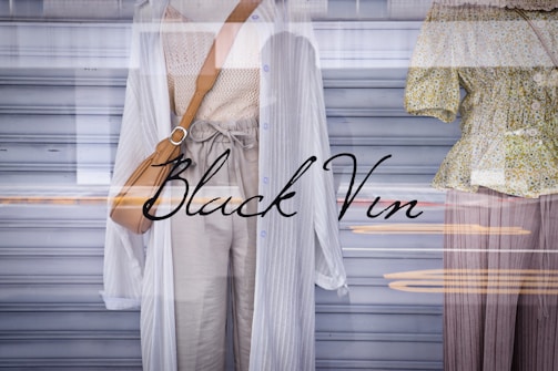 Two mannequins in a store window display are dressed in light, casual clothing. One wears a long white cardigan over a beige knitted top and light trousers, complemented by a tan shoulder bag. The other mannequin is dressed in a green floral blouse with buttons and wide brown trousers. The display reflects urban street scenery in the glass, adding layers to the image.