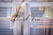 Two mannequins in a store window display are dressed in light, casual clothing. One wears a long white cardigan over a beige knitted top and light trousers, complemented by a tan shoulder bag. The other mannequin is dressed in a green floral blouse with buttons and wide brown trousers. The display reflects urban street scenery in the glass, adding layers to the image.