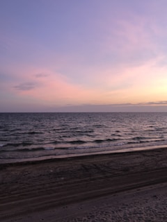 Soft pastel colors of the sky reflect on the calm sea, creating a serene atmosphere. Gentle waves lap against a sandy beach, with a subtle gradient from sand to sea.