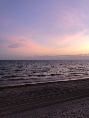 Soft pastel colors of the sky reflect on the calm sea, creating a serene atmosphere. Gentle waves lap against a sandy beach, with a subtle gradient from sand to sea.