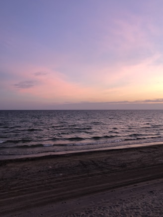 Soft pastel colors of the sky reflect on the calm sea, creating a serene atmosphere. Gentle waves lap against a sandy beach, with a subtle gradient from sand to sea.