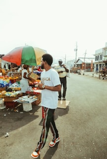 A man wearing a white t-shirt and patterned pants is walking by a street market. Beside him, under a colorful umbrella, a vendor arranges goods displayed on a table. Various items, such as food packets and beverages, are available for sale. Another man wearing a cap is in the background, adding to the bustling street scene.