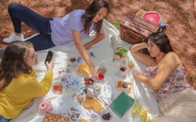 Three people enjoying a picnic on a white blanket surrounded by pine needles. There is a variety of food and drinks including berries, cookies, and tea. A wicker picnic basket and a pink instant camera are also visible. The scene includes playful details such as Polaroid photos scattered around, creating a casual and relaxed atmosphere.