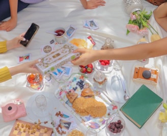 A picnic setting on a white blanket with people sitting around. The center features various snacks like berries, pastries, and cookies on a floral tray. A chessboard with scattered pieces lies nearby. Hands are exchanging a decorative box, and there is also a smartphone, a green book, cameras, playing cards, and a vase with pink flowers.