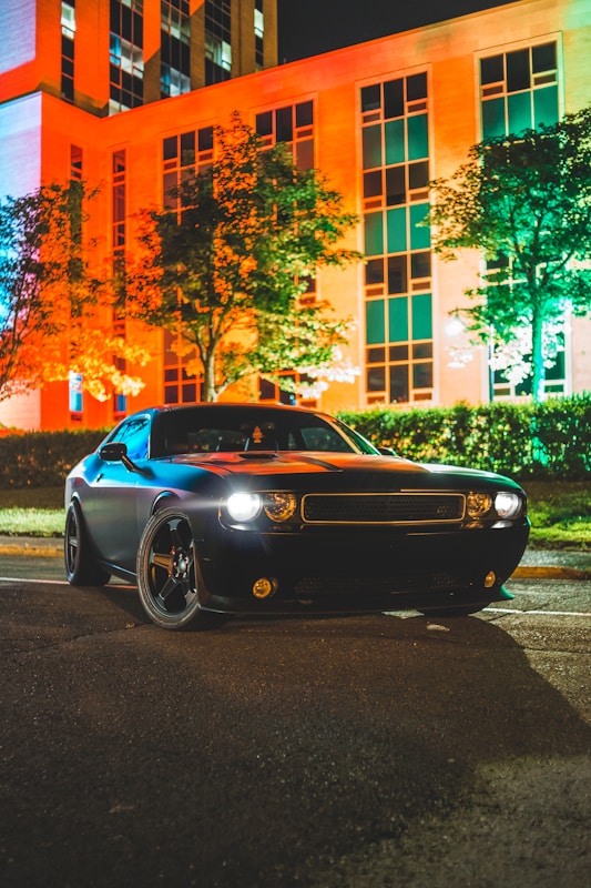 A sleek black car is parked on a street at night, with illuminated headlights casting light on the ground. The background features a tall building with large windows, where vibrant orange and teal lights highlight the facade and nearby trees.