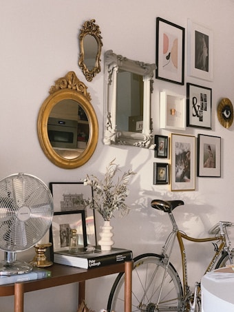 A corner of a room featuring a wall adorned with various framed artwork, including paintings and mirrors. The wall has an eclectic mix of frames, sizes, and styles. Below the wall is a wooden shelf displaying a metallic table fan, books, and a vase with dried flowers. Also, a vintage bicycle is partially visible, leaning against the wall.