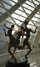 Three bronze sculptures of dancing figures with elongated limbs and contemporary attire are positioned in an indoor space with large glass windows. Sunlight streams in, creating dynamic shadows and reflections on the polished floor. The backdrop features modern architectural elements and glimpses of trees.