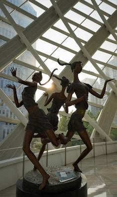 Three bronze sculptures of dancing figures with elongated limbs and contemporary attire are positioned in an indoor space with large glass windows. Sunlight streams in, creating dynamic shadows and reflections on the polished floor. The backdrop features modern architectural elements and glimpses of trees.