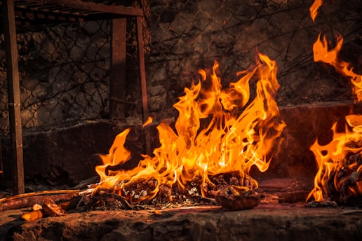 Vivid flames in shades of orange and yellow consume a pile of materials on a stone surface, with metal and chain-link fencing visible in the background.