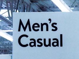 A signage featuring bold, black text reads 'Men's Casual' against a light background. The setting appears to be indoors, with visible overhead lighting and structural elements like beams creating an industrial look.
