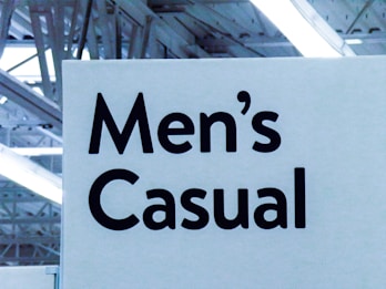 A signage featuring bold, black text reads 'Men's Casual' against a light background. The setting appears to be indoors, with visible overhead lighting and structural elements like beams creating an industrial look.