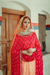 A person is dressed in traditional attire featuring a bright red and orange embellished outfit. The outfit includes a dupatta draped over the head, adorned with intricate embroidery and beadwork. Jewelry including a large necklace, earrings, and a nose ring complements the ensemble. The background consists of a rustic, partially painted wall with a wooden door.