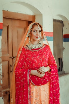 A person is dressed in traditional attire featuring a bright red and orange embellished outfit. The outfit includes a dupatta draped over the head, adorned with intricate embroidery and beadwork. Jewelry including a large necklace, earrings, and a nose ring complements the ensemble. The background consists of a rustic, partially painted wall with a wooden door.