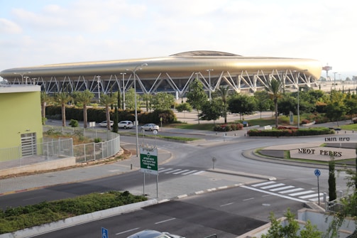 A large, modern stadium with a golden hue dominates the scene, supported by numerous structural beams. Surrounding the stadium are lush green trees and well-maintained landscaping, including palm trees. The area features clear roads, a roundabout, and a variety of signs. In the foreground, there's a building with a pale green facade.