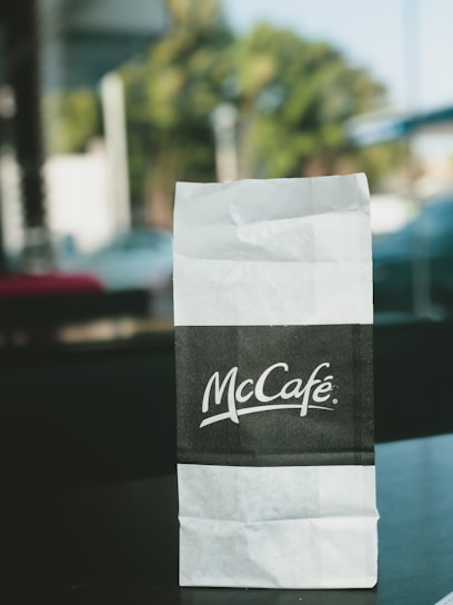 A McCafé branded paper bag is placed on a table with a blurred outdoor background. The bag is predominantly white with a black section displaying the McCafé logo in white text.
