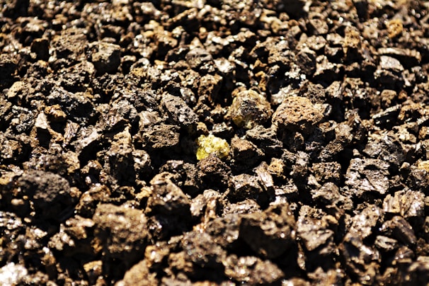 A close-up view of a surface covered with rough, jagged pieces of dark brown volcanic rock, with a few shiny yellowish minerals visible among the stones.