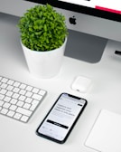 A modern workspace setup featuring an iMac with a visible Apple logo, a white keyboard, and a trackpad. An iPhone displaying an app interface lies next to an AirPods case. A small, round green plant in a white pot adds a touch of nature to the minimalist scene.