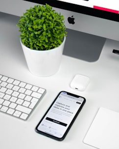 A modern workspace setup featuring an iMac with a visible Apple logo, a white keyboard, and a trackpad. An iPhone displaying an app interface lies next to an AirPods case. A small, round green plant in a white pot adds a touch of nature to the minimalist scene.