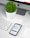 A modern workspace setup featuring an iMac with a visible Apple logo, a white keyboard, and a trackpad. An iPhone displaying an app interface lies next to an AirPods case. A small, round green plant in a white pot adds a touch of nature to the minimalist scene.