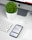 A modern workspace setup featuring an iMac with a visible Apple logo, a white keyboard, and a trackpad. An iPhone displaying an app interface lies next to an AirPods case. A small, round green plant in a white pot adds a touch of nature to the minimalist scene.