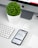 A modern workspace setup featuring an iMac with a visible Apple logo, a white keyboard, and a trackpad. An iPhone displaying an app interface lies next to an AirPods case. A small, round green plant in a white pot adds a touch of nature to the minimalist scene.