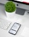 A modern workspace setup featuring an iMac with a visible Apple logo, a white keyboard, and a trackpad. An iPhone displaying an app interface lies next to an AirPods case. A small, round green plant in a white pot adds a touch of nature to the minimalist scene.