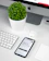 A modern workspace setup featuring an iMac with a visible Apple logo, a white keyboard, and a trackpad. An iPhone displaying an app interface lies next to an AirPods case. A small, round green plant in a white pot adds a touch of nature to the minimalist scene.