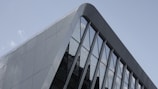 A section of a modern building facade featuring sleek, angular metallic panels and large reflective glass windows. The architecture is minimalist and geometric, with a blend of sharp and curved lines.