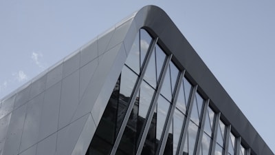 A section of a modern building facade featuring sleek, angular metallic panels and large reflective glass windows. The architecture is minimalist and geometric, with a blend of sharp and curved lines.