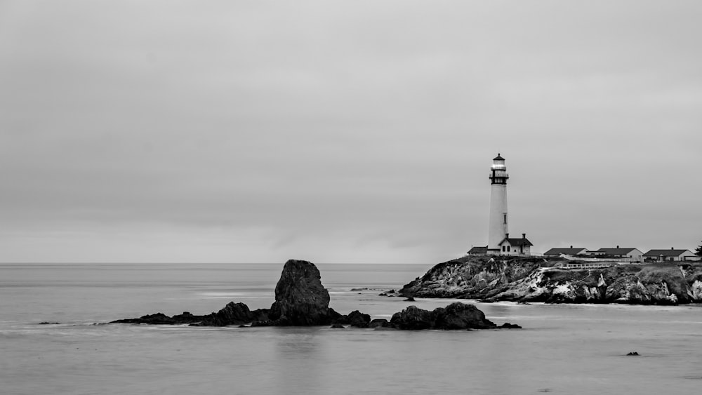 Foto en escala de grises del faro en una formación rocosa cerca del cuerpo de agua