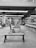 A gym area with rows of weight benches and racks filled with dumbbells. The room is spacious and well-lit with ceiling lights. Mirrors line the walls, reflecting the equipment and creating a sense of depth. The floor is covered with rubber mats.