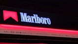 A signboard with the word 'Marlboro' illuminated in white against a dark background, accompanied by a red geometric shape resembling a chevron above the text. Below the main sign, there's text in Japanese, which appears to be a warning or disclaimer.