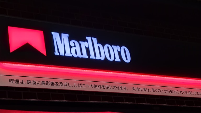 A signboard with the word 'Marlboro' illuminated in white against a dark background, accompanied by a red geometric shape resembling a chevron above the text. Below the main sign, there's text in Japanese, which appears to be a warning or disclaimer.