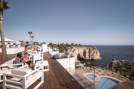 A seaside resort with people relaxing on a wooden deck featuring white cushioned furniture and tables. In the background, the ocean is visible along with a rocky cliff and clear sky. The setting appears leisurely, with palm trees dotting the area.