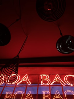 Bright red neon lights spell out the words against a dark background with black circular metal objects hanging from the ceiling, creating a bold, modern, and industrial aesthetic.