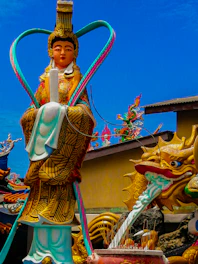 A colorful religious or cultural statue featuring intricate details and vibrant designs. The statue depicts a deity or figure adorned in elaborate traditional attire, standing alongside a dragon whose mouth emits a stream. Incense sticks are placed in a holder at the base in front of the statue.