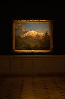 A framed painting of a mountain landscape is displayed on a dark wall. The artwork features snow-capped peaks under a partly cloudy sky, with lush greenery in the foreground.