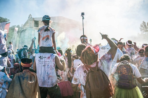 A lively crowd is participating in a colorful outdoor event. People are wearing white shirts stained with various vibrant powders. In the center, a person is carrying a child on their shoulders, and others are raising their hands and holding cameras. Banners and flags are visible in the background, with a festive atmosphere surrounding the scene.