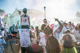 A lively crowd is participating in a colorful outdoor event. People are wearing white shirts stained with various vibrant powders. In the center, a person is carrying a child on their shoulders, and others are raising their hands and holding cameras. Banners and flags are visible in the background, with a festive atmosphere surrounding the scene.