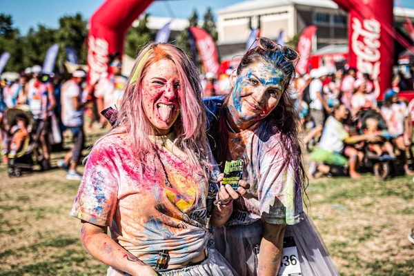 Two people are covered in vibrant colored powder, smiling and enjoying themselves in a lively outdoor event setting. The background features other participants, event banners, and greenery.