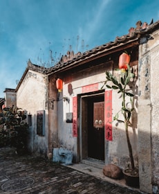 A weathered, traditional Chinese building with red lanterns hanging by the entrance. The walls are adorned with red banners containing Chinese characters. The old brick pathway leads to the doorway, surrounded by potted plants and casting shadows from the sunlight.