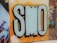 Bold graffiti art featuring large, stylized letters 'SMO' painted in pastel blue with a dark outline against an orange and yellow gradient background. The surrounding wall has additional graffiti with various colors and designs. The phrase 'smile more often...' is written in white on the left, adding a positive note.