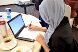 A person wearing a gray headscarf is seated at a round table, engaged in writing on paper placed over a laptop. The table is scattered with various items including a cup of tea, a plate with food remnants, a reusable coffee cup, and two bottles of water. A projector is visible in the background.