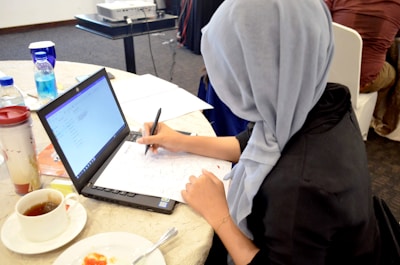 A person wearing a gray headscarf is seated at a round table, engaged in writing on paper placed over a laptop. The table is scattered with various items including a cup of tea, a plate with food remnants, a reusable coffee cup, and two bottles of water. A projector is visible in the background.