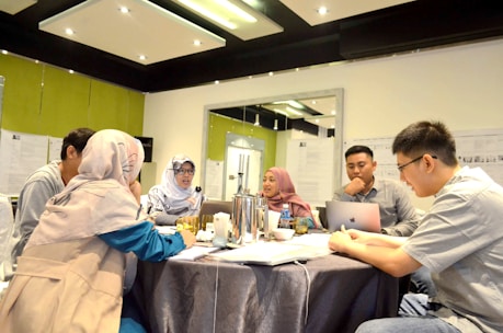 A group of five people are gathered around a table in a meeting room, discussing and working together. Two people are wearing headscarves, and one person is using a laptop. The room is well-lit, with papers and documents on the walls, and there is a silver thermos and drinks on the table.