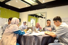 A group of five people are gathered around a table in a meeting room, discussing and working together. Two people are wearing headscarves, and one person is using a laptop. The room is well-lit, with papers and documents on the walls, and there is a silver thermos and drinks on the table.