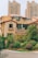 A residential area featuring a house with a beige facade and reddish-brown roof tiles, surrounded by lush greenery and shrubs. In the background, there are tall, modern apartment buildings with numerous windows and balconies, contributing to an urban yet peaceful setting.
