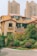 A residential area featuring a house with a beige facade and reddish-brown roof tiles, surrounded by lush greenery and shrubs. In the background, there are tall, modern apartment buildings with numerous windows and balconies, contributing to an urban yet peaceful setting.